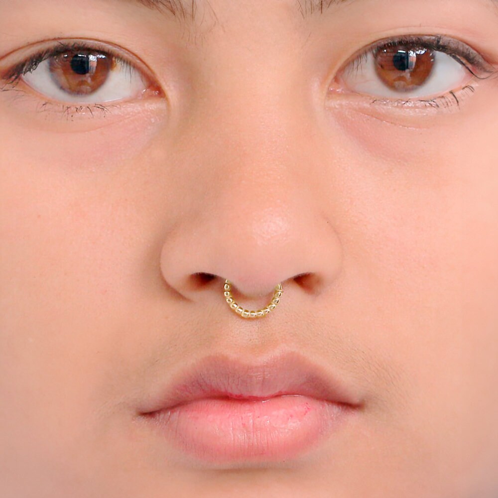 Close-up of a person's face, focusing on their nose with the Lysa Elegant Design Septum from Kaya Movement. The person's skin is smooth, and their eyes are brown. The gold septum ring features a delicate chain design and adds a subtle shine for everyday elegance. The lips are slightly parted, and minimal makeup is worn.